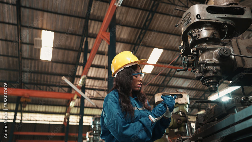 Fototapeta premium African factory worker in safety wear is checking a piece of industrial metal while Caucasian coworker help her to combine metal part to machine. Two production engineers are working in metal factory.
