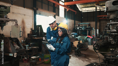 Two multiracial factory workers in safety wear are using a tablet to plan for working at a metalwork production factory. Male and female production engineers are examining the status of manufacturing.