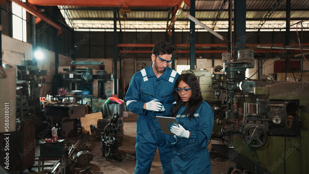 Two multiracial factory workers in safety wear are using a tablet to plan for working at a metalwork production factory. Male and female production engineers are examining the status of manufacturing. Two multiracial factory workers in safety wear are using a tablet to plan for working at a metalwork production factory. Male and female production engineers are examining the status of manufacturing.