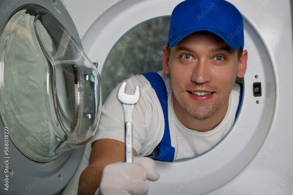 Positive plumber looks out of an open hole for loading laundry into washing machine with wrench in his hand. Young master in blue robe and a hat advertises high-quality repair of household appliances Positive plumber looks out of an open hole for loading laundry into washing machine with wrench in his hand. Young master in blue robe and a hat advertises high-quality repair of household appliances