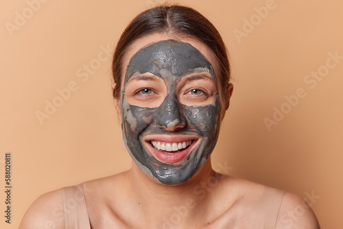 Horizontal shot of beautiful brunette European woman smiles broadly applies nourishing clay mask enjoys healing facial treatment stands against brown background want to look fresh and young.