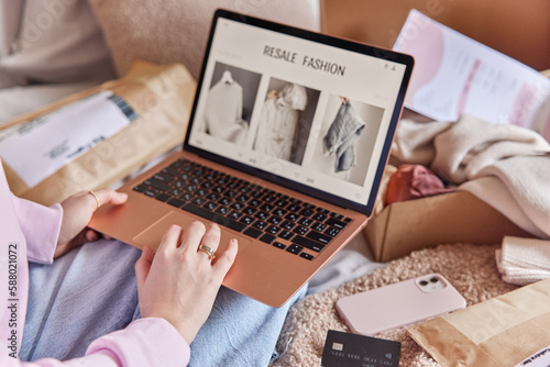 Unrecognizable woman uses laptop computer for shopping online searches clothes to buy on resale fashion site surrounded by smartphone credit card and paper packages with clothes makes purchases photo