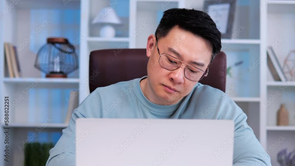Bored, sad, exhausted Asian male office worker with glasses is sitting at desk in office, trying to work on his laptop. Man shows no interest in his work and appears to be suffering from burnout. Bored, sad, exhausted Asian male office worker with glasses is sitting at desk in office, trying to work on his laptop. Man shows no interest in his work and appears to be suffering from burnout.