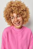 Vertical shot of positive carefree young woman with bushy curly hair looks dreamily aside laughs at something dressed in pink pullover optical eyeglasses poses indoors shows genuine happiness