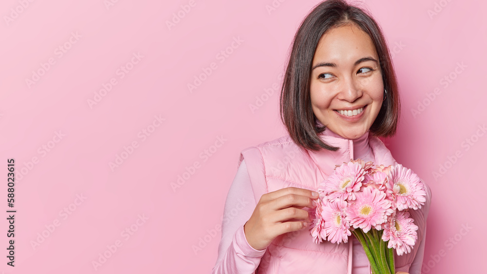 Horizontal shot of beautiful glad woman holds bouquet of gerbera flowers got from beloved person smiles gently focused aside wears vest isolated over pink studio background copy space for your advert
