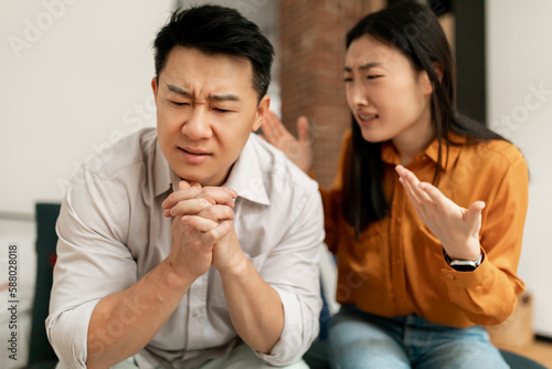 Angry asian lady shouting at her middle aged husband, having conflict while sitting on sofa at home, focus on upset man