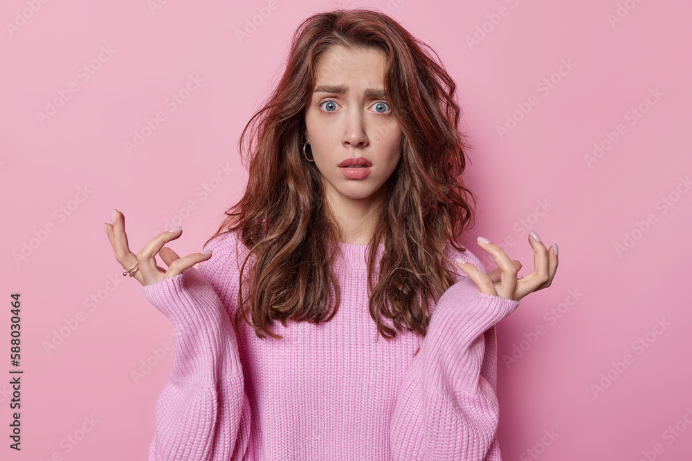 Confused hesitant European woman with dark wavy hair spreads palms feels unaware dressed in knitted jumper stands indignant faces dilemma isolated over pink background. Human reactions concept Confused hesitant European woman with dark wavy hair spreads palms feels unaware dressed in knitted jumper stands indignant faces dilemma isolated over pink background. Human reactions concept