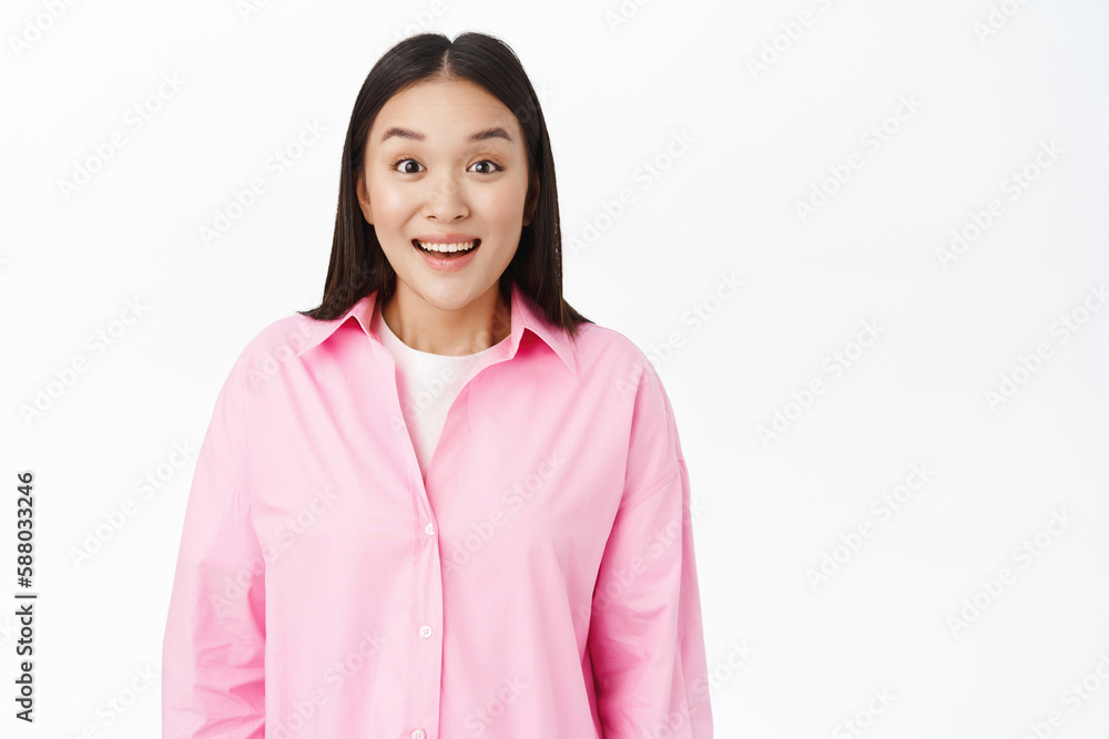 Smiling korean girl looking excited and surprised at camera, amazed by smth, standing over white studio background Smiling korean girl looking excited and surprised at camera, amazed by smth, standing over white studio background