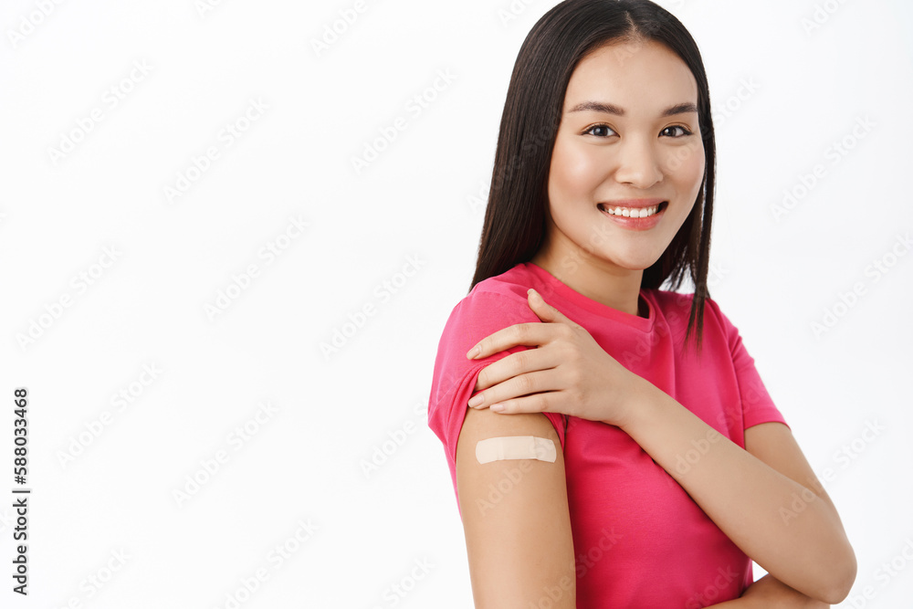 Vaccination from covid. Smiling asian girl shows her shoulder with patch after coronavirus vaccine shot, looking happy, standing over white background Vaccination from covid. Smiling asian girl shows her shoulder with patch after coronavirus vaccine shot, looking happy, standing over white background
