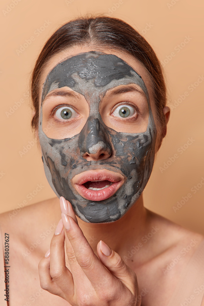 Headshot of shocked stunned woman with dark combed hair gets beauty treatments applies facial clay mask holds breath from amazement poses indoor takes care of skin isolated over brown background Headshot of shocked stunned woman with dark combed hair gets beauty treatments applies facial clay mask holds breath from amazement poses indoor takes care of skin isolated over brown background