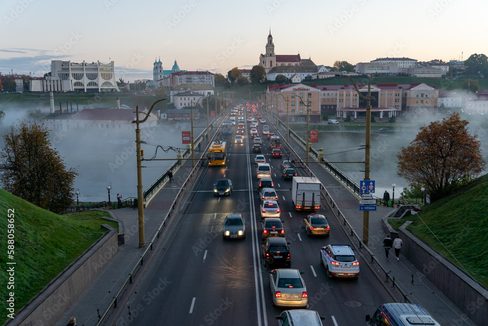 Obraz premium Belarus, Grodno July 15, 2022: The movement of cars and public transport on the bridge over the river. Thick morning fog over the bridge in the city. fog over a crowded road