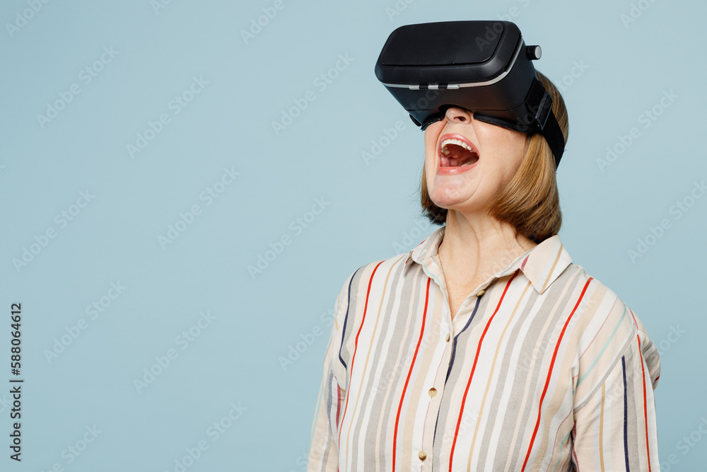 Elderly surprised shocked amazed woman 50s years old wear striped shirt watching in vr headset pc gadget isolated on plain pastel light blue cyan color background studio portrait. Lifestyle concept. Elderly surprised shocked amazed woman 50s years old wear striped shirt watching in vr headset pc gadget isolated on plain pastel light blue cyan color background studio portrait. Lifestyle concept.