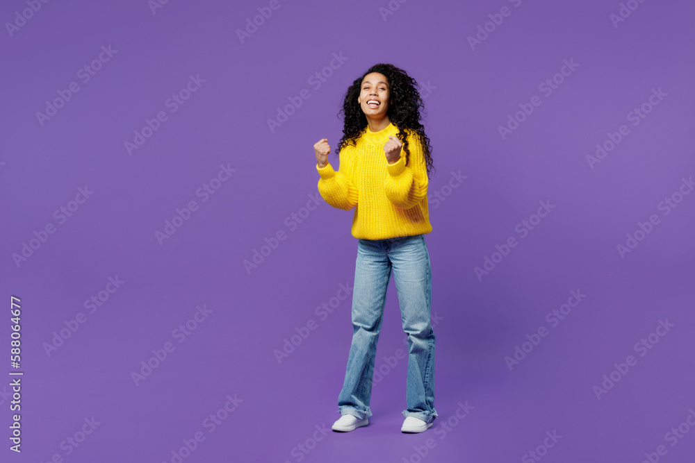 Full body side profile view happy young woman of African American ethnicity wears casual yellow sweater doing winner gesture clench fist isolated on plain purple background studio Lifestyle concept. Full body side profile view happy young woman of African American ethnicity wears casual yellow sweater doing winner gesture clench fist isolated on plain purple background studio Lifestyle concept.