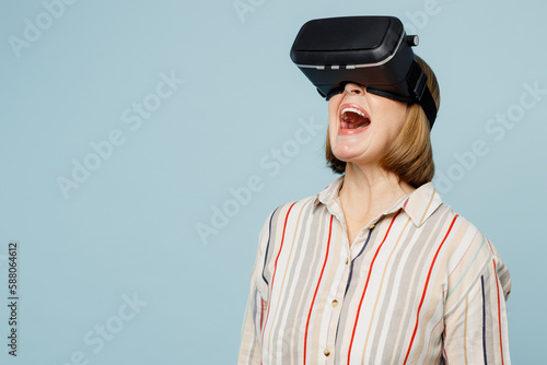 Elderly surprised shocked amazed woman 50s years old wear striped shirt watching in vr headset pc gadget isolated on plain pastel light blue cyan color background studio portrait. Lifestyle concept. © ViDi Studio