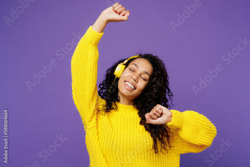 Young smiling fun woman of African American ethnicity wear casual yellow sweater headphones listen to music dance raise up hands isolated on plain purple background studio portrait. Lifestyle concept.