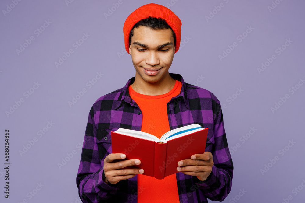 Young smiling smart clever student man of African American ethnicity wear casual shirt orange hat reading book novel at library isolated on plain pastel light purple color background studio portrait. Young smiling smart clever student man of African American ethnicity wear casual shirt orange hat reading book novel at library isolated on plain pastel light purple color background studio portrait.