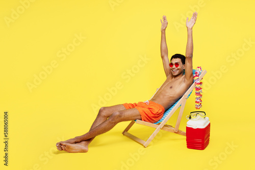 Full body side view fun young sexy man wear orange shorts swimsuit relax near hotel pool sit in deckchair raise up hands isolated on plain yellow background. Summer vacation sea rest sun tan concept.