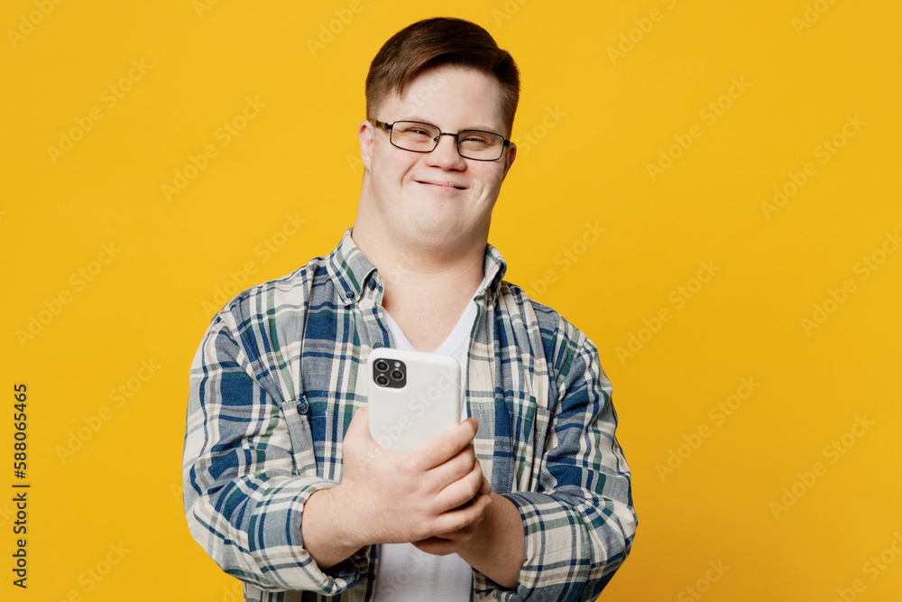 Young satisfied fun smiling man with down syndrome wears glasses casual clothes hold in hand use mobile cell phone isolated on pastel plain yellow color background. Genetic disease world day concept. Young satisfied fun smiling man with down syndrome wears glasses casual clothes hold in hand use mobile cell phone isolated on pastel plain yellow color background. Genetic disease world day concept.