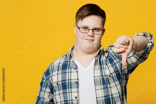 Young sad displeased dissatisfied man with down syndrome wear glasses casual clothes showing thumb down dislike gesture isolated on pastel plain yellow background. Genetic disease world day concept.