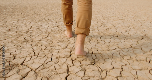 Close up shot of feet of adult man walking barefoot on bottom of dried lake or river, stepping on cracked soil ground destroyed by erosions - ecological issues concept 