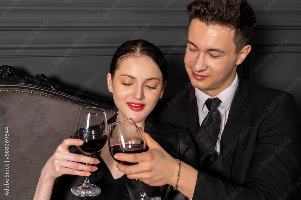 A young couple elegantly and stylishly dressed spend a romantic time with glasses of wine in hand while sitting on a vintage couch in a beautiful dark room in Gothic style. A romantic date.