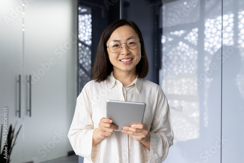 Portrait of successful asian female programmer inside office, young woman smiling and looking at camera, using tablet computer to test application software, workplace inside office.