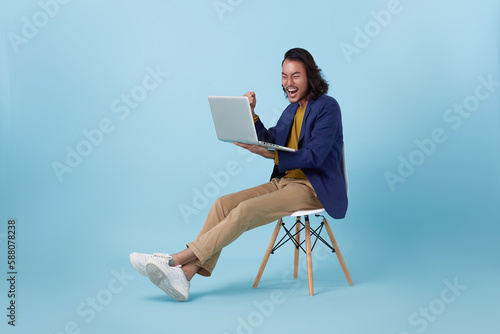 happy celebration asian business man achievement goal using computer laptop sitting on white chair isolated on blue studio background..