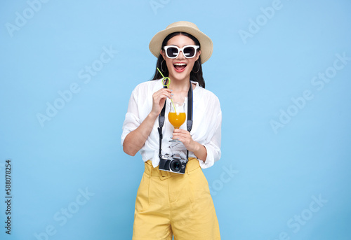 Happy smiling young asian woman tourist in summer hat standing with camera and drinking orange juice isolated on blue studio background