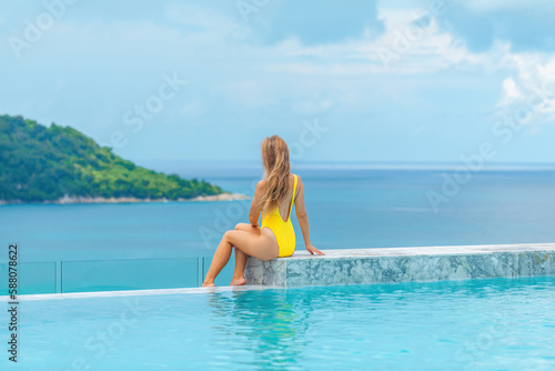 Back view of young woman in yellow swimwear relaxes in infinity pool at a luxury spa resort, enjoying the stunning sea view. A perfect summer vacation getaway.