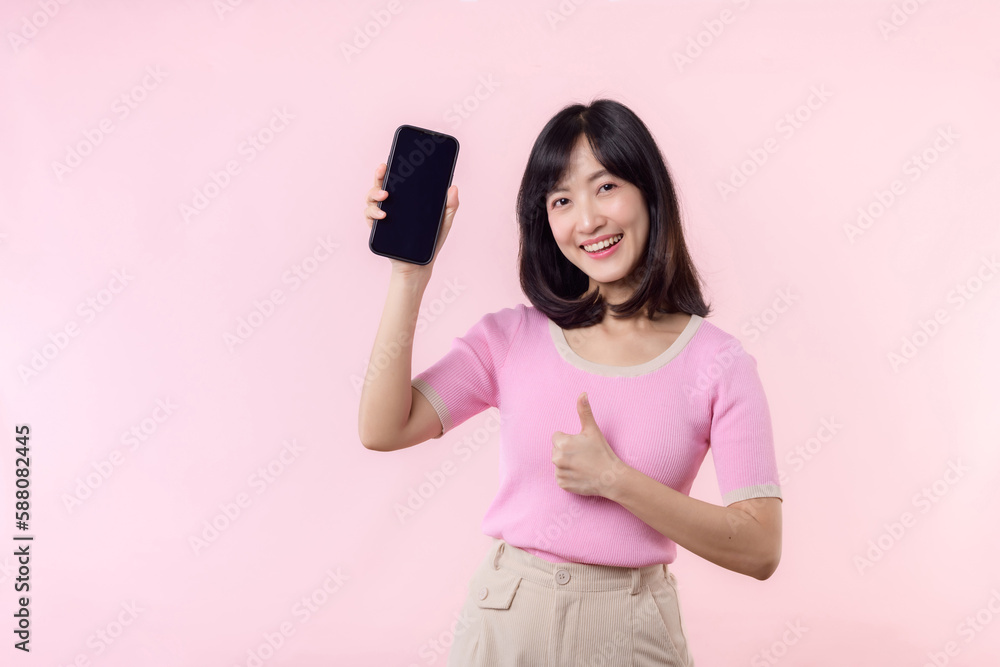 Portrait beautiful young asian woman happy smile showing smartphone screen with pointing finger hand gesture to mobile phone isolated on pink pastel studio background. Blank screen smartphone concept