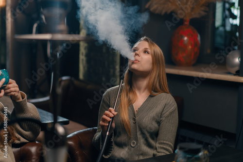 Beautiful young woman inhaling hookah girl smoking hookah sitting in lounge bar concept of relaxation and smoking