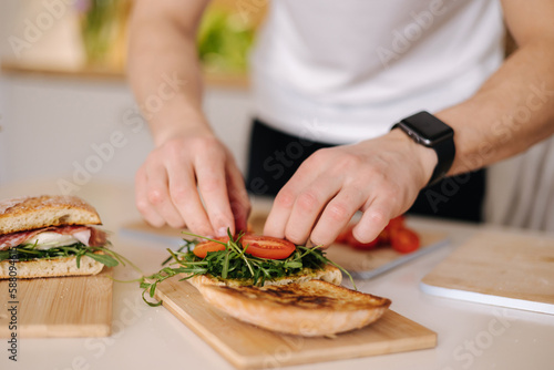 Young man put tomatoes on sandwich. Man preparing lunch at home