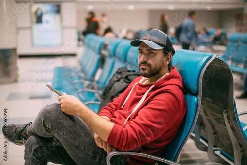 A young, handsome man is sitting with a phone in his hands at the airport in the evening. A guy, a tourist with a smartphone and a backpack is waiting for departure at the gate © Anna