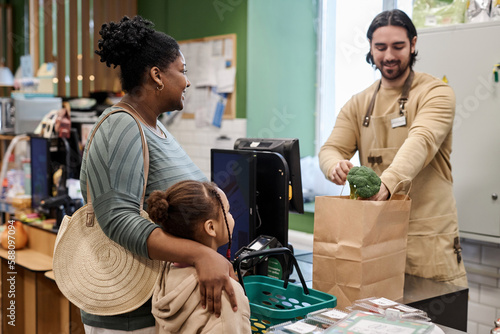 Portrait of male cashier in supermarket helping young mother with groceries and packing vegetables to paper bag photo
