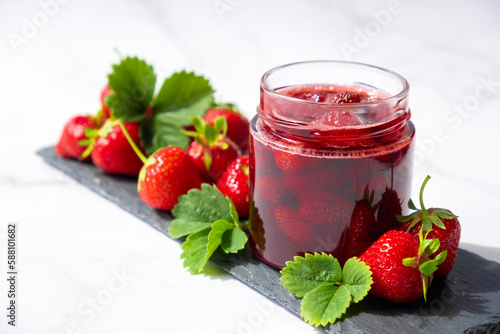 Strawberry jam in glass jar on slate dish with fresh strawberry fruit and green leaves on marble. Recipe of delicious berry jam of strawberry full of vitamins and antioxidants.