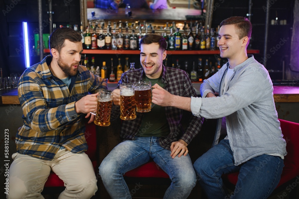 Obraz premium Three young men in casual clothes are smiling and clanging glasses of beer together while sitting at bar counter in pub
