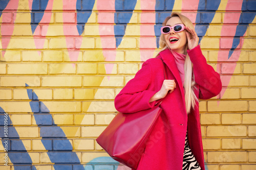 Fotomurale Fashionable happy smiling blonde woman wearing trendy pink sunglasses, fuchsia color coat, with faux leather tote, shopper bag, posing on colorful background