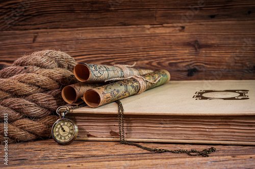 Old items, map scrolls and a pocket watch lying on an old book next to a thick rope