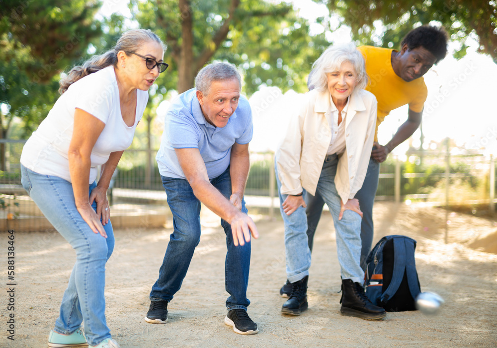 A group of multiracial middle aged mixed-sex adult people playing petanque game outdoors in public park on a sunny day