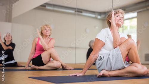 Smiling senior woman doing yoga with group in fitness studio, sitting in twisting asana Matsyendrasana .