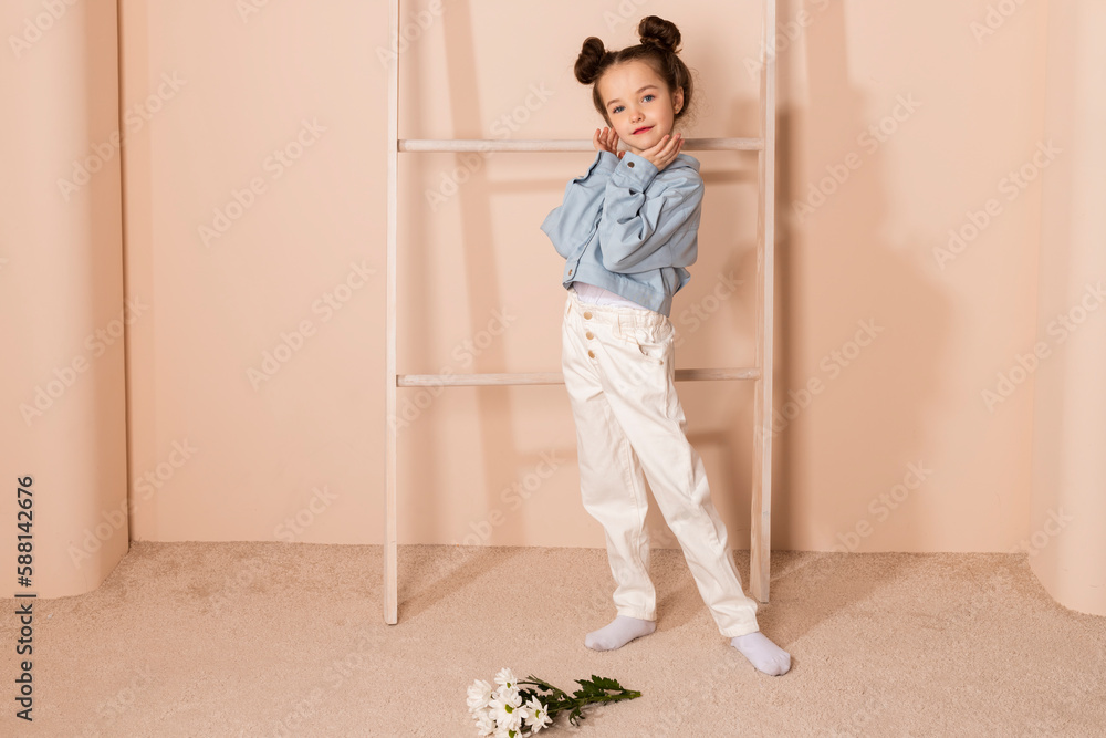 Portrait of a charming little girl, well dressed in a blue shirt and white pants or trousers and with a slight makeup and double bun hair. Isolated on pastel beige background. Portrait of a charming little girl, well dressed in a blue shirt and white pants or trousers and with a slight makeup and double bun hair. Isolated on pastel beige background.