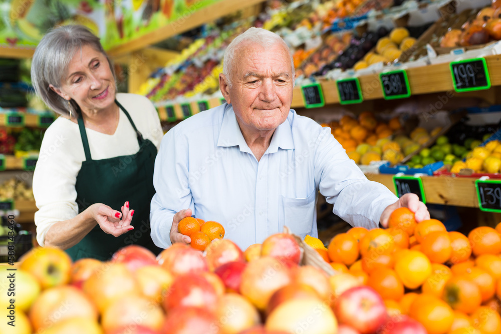 Old man choosing mandarins in greengrocer. Mature woman worker helping him with it.