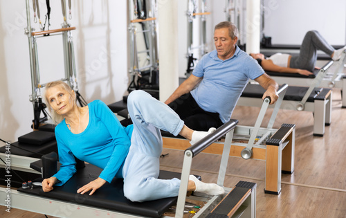 Focused elderly woman doing toning exercise for back on Pilates reformer bed in health fitness center