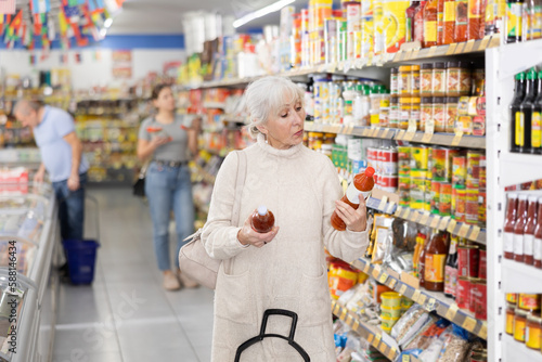 Focused interested gray-haired elderly woman standing near shelves with condiments in supermarket, reading labels on bottles with sauce..
