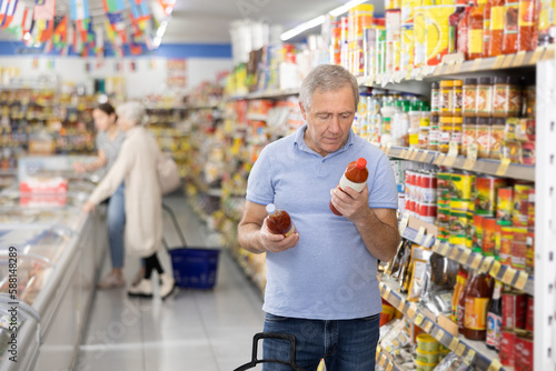 Focused aged man reading labels on bottles with sauces in supermarket, carefully examining ingredients and expiration date while shopping for groceries