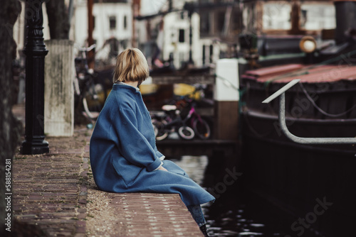 An unrecognizable woman in a stylish blue coat is sitting on the pier. Residential area in the Netherlands. Toned.