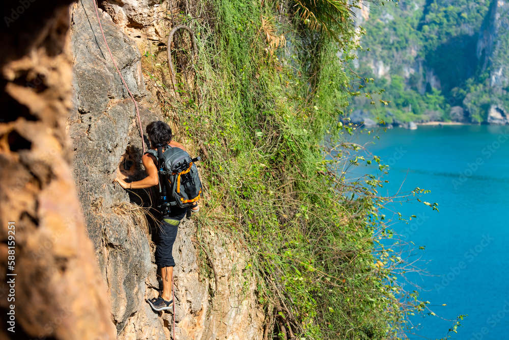 Obraz premium Young Asian man climber climbing on rocky coastline at tropical island in sunny day. Handsome guy enjoy outdoor active lifestyle and extreme sport training mountain climbing on summer holiday vacation
