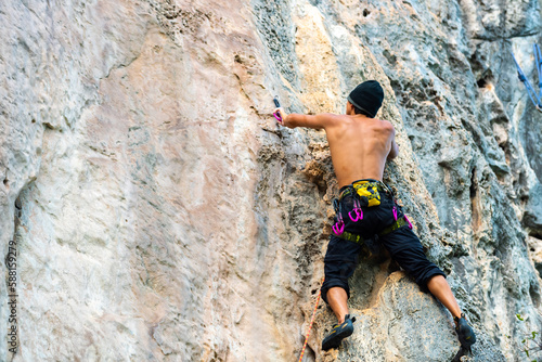 Young Asian man climber climbing on rocky coastline at tropical island in sunny day. Handsome guy enjoy outdoor active lifestyle and extreme sport training mountain climbing on summer holiday vacation