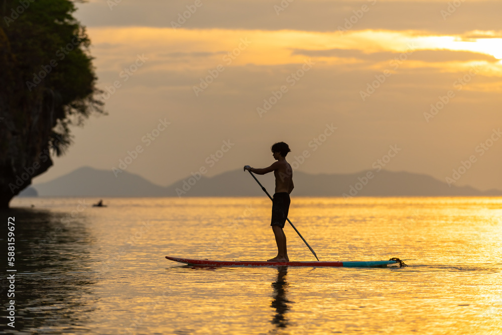 Young Asian man standing on paddle board and rowing in the ocean at tropical island at sunset. Wellness man enjoy outdoor lifestyle and water sports surfing and paddle boarding on summer vacation.