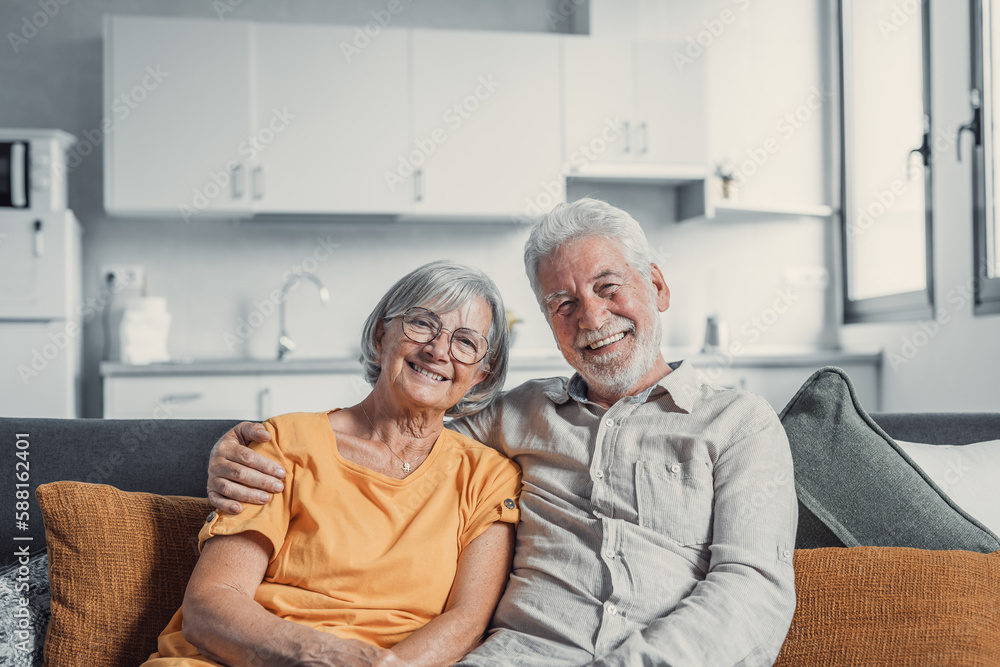 Headshot portrait of smiling elderly 60s husband and wife sit relax on couch hugging cuddling, happy mature old couple rest on sofa in living room embrace look at camera show love and care. Headshot portrait of smiling elderly 60s husband and wife sit relax on couch hugging cuddling, happy mature old couple rest on sofa in living room embrace look at camera show love and care.